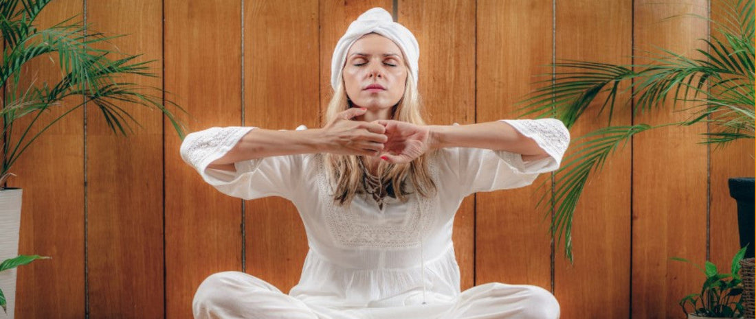 Egy fehér ruhás hölgy kundalini jógapózt végez egy természetes közegben.