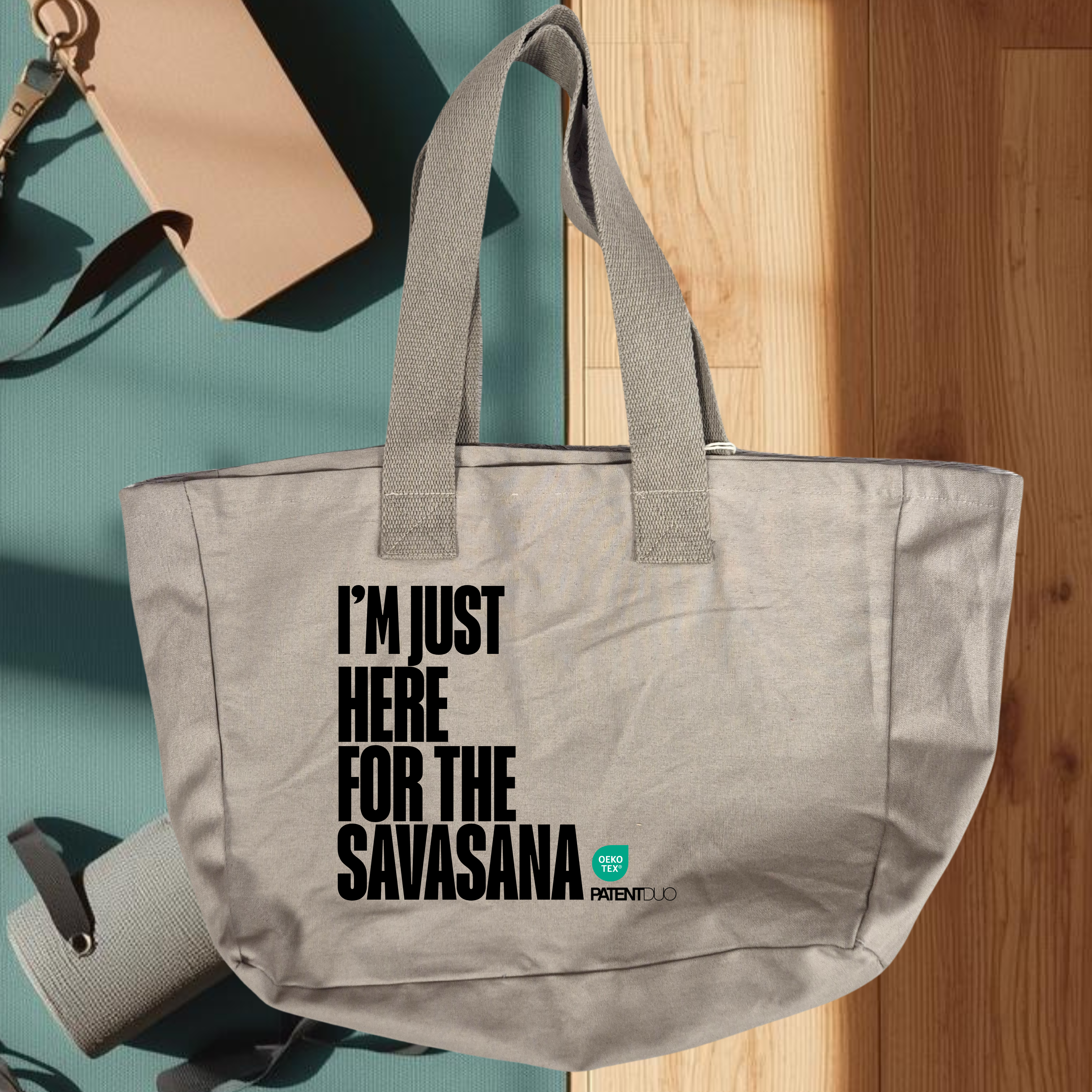 A PatentDuo I'M JUST HERE FOR THE SAVASANA - Prémium pamut shopper egy világosszürke, nagy kapacitású szállítmány merész fekete szöveggel és egy kis zöld logóval, amely egy jógamatracon látható fitness kiegészítők között. szürke, gray színben