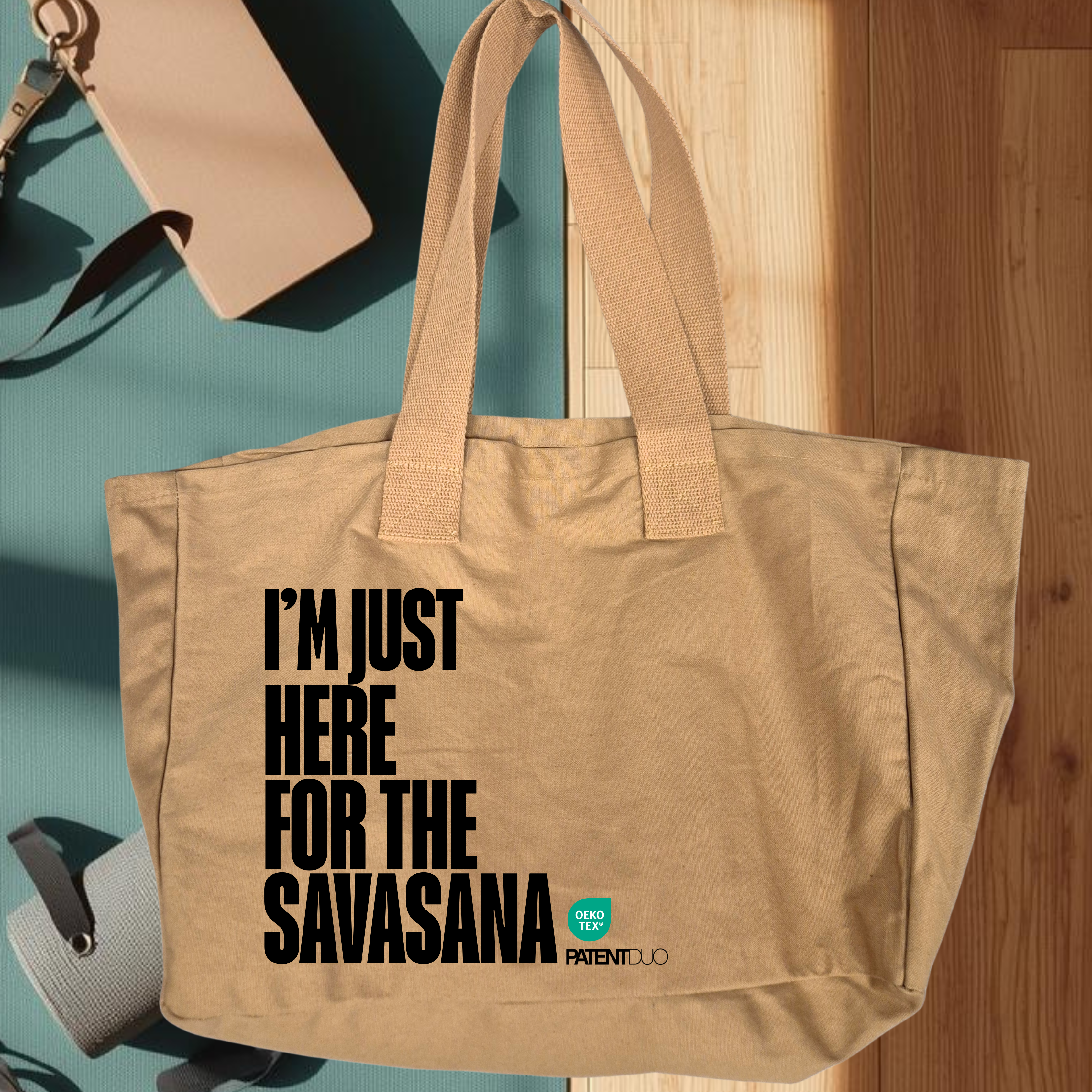 A PatentDuo I'M JUST HERE FOR THE SAVASANA - prémium pamut shopper egy jógaszőnyegen pihen, mellette kulacs, telefon és jógaheveder látható, miközben napsütés árad a fapadlóra. mokka szín