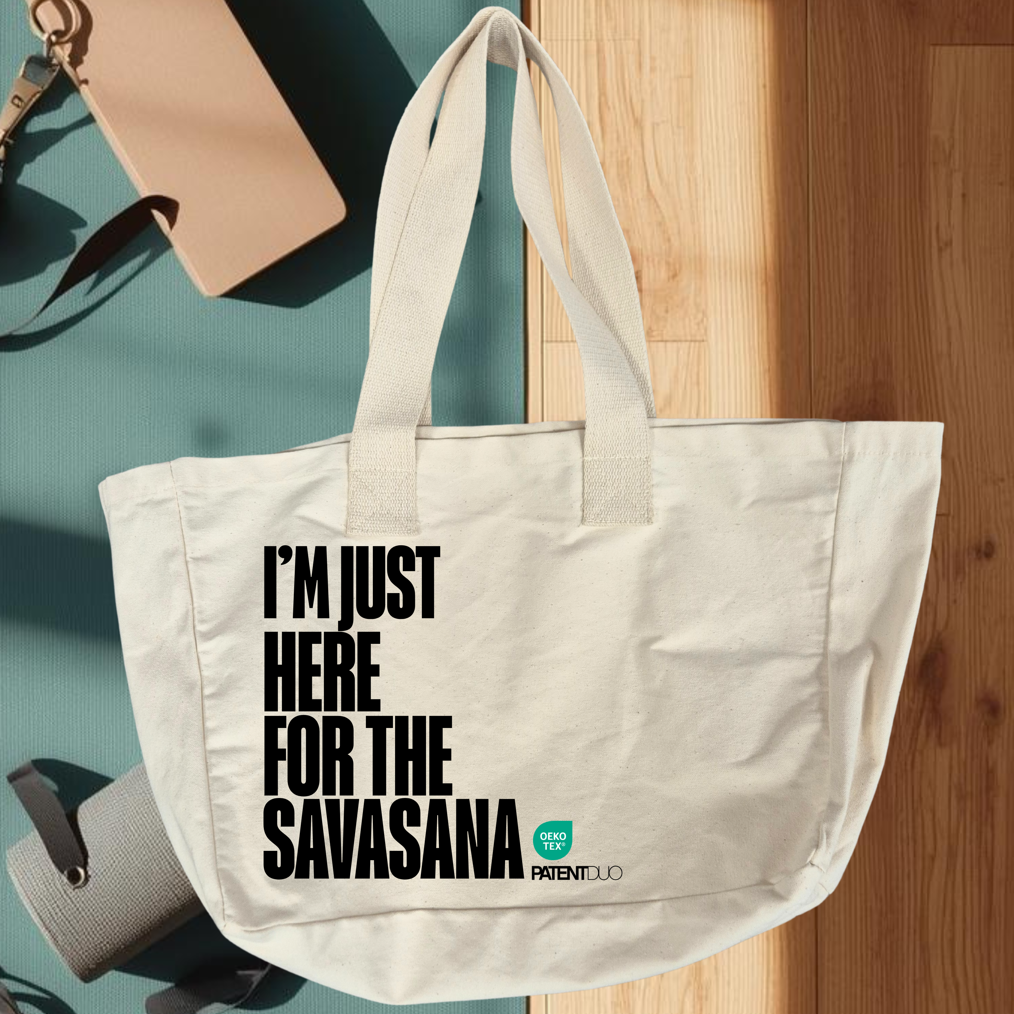 Az I'M JUST HERE FOR THE SAVASANA - Prémium pamut shopper (PatentDuo) fehér színben, nagy fekete felirattal, kék jógaszőnyegen pihen napsütötte fapadlón, beige kulacs és feltekert jógamatrac mellett. natur színben